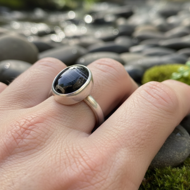 Silver ring with a black stone on a hand against a natural background of stones and water.