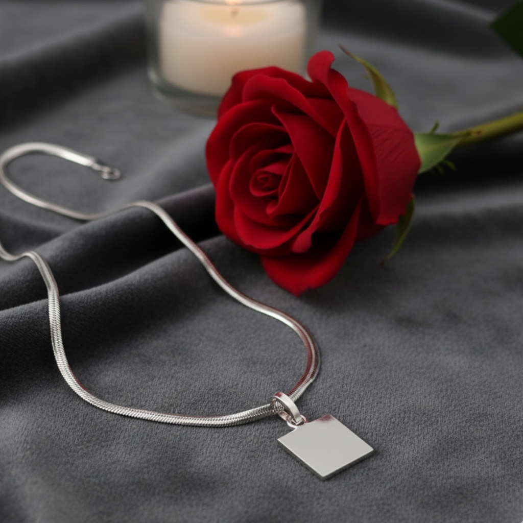 Silver necklace with a square pendant next to a red rose on gray fabric with a candle in the background